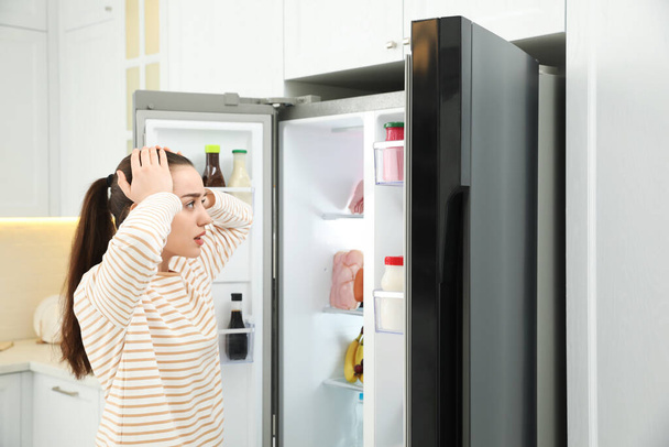 Emotional young woman near open refrigerator in kitchen - Fotografie, Obrázek