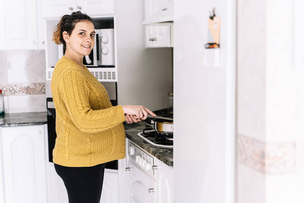 Schwangere mit gelbem Pullover steht in einer Küche neben einem Kühlschrank, lächelt und dreht das Gesicht nach rechts - Foto, Bild