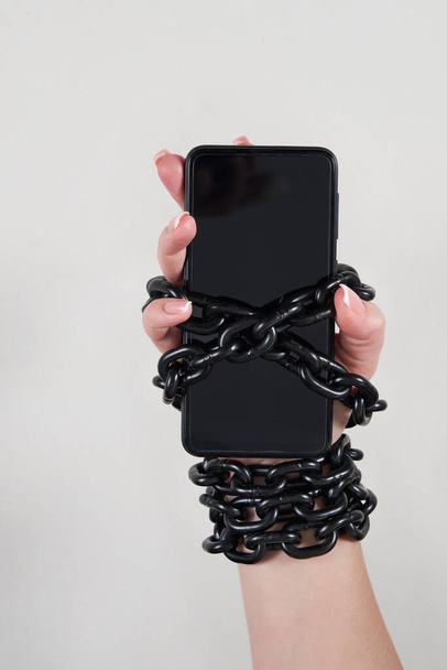 Cadena de hierro que une mano y smartphone en concepto de redes sociales y adicción a Internet sobre fondo gris - Foto, imagen