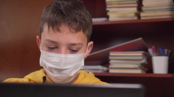 Okul malzemeleriyle dolu ahşap bir dolabın arka planında koruyucu bir yüz maskesi takmış bir dizüstü bilgisayarla evde çalışan bir genç. Koronavirüs karantinası sırasında - Video, Çekim