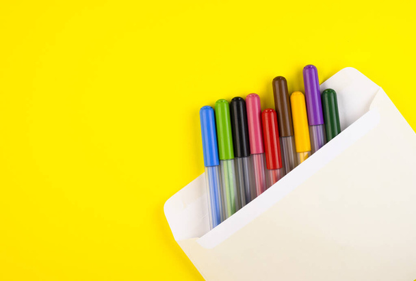 markers of different colors are enclosed in a mail envelope on a yellow background, top view. markers close up laid out in a row - Фото, изображение