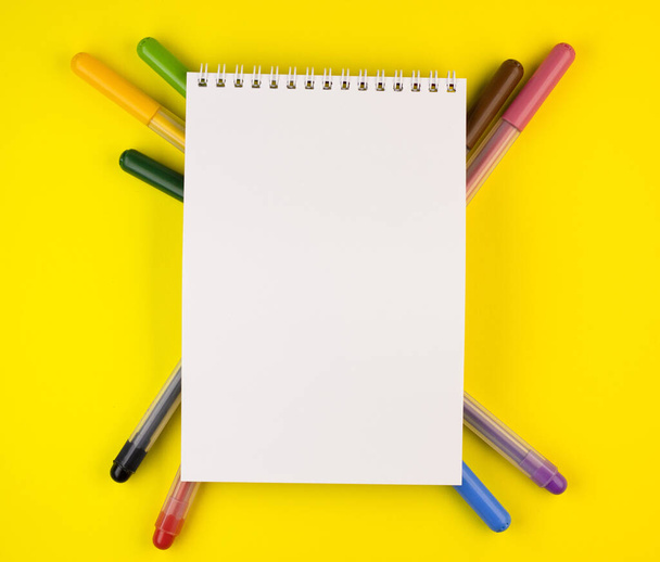 markers of different colors with a white Notepad on a yellow background, top view. colorful pens are spread out under the notebook close up - Photo, Image