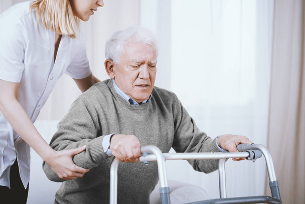 Старший серый мужчина с ходунком и молодой медсестрой, поддерживающей его
 - Фото, изображение
