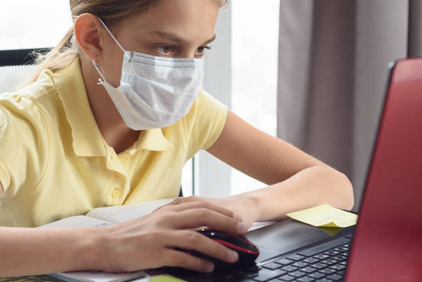 Ένα κορίτσι με ιατρική μάσκα σπουδάζει στον υπολογιστή.. - Φωτογραφία, εικόνα