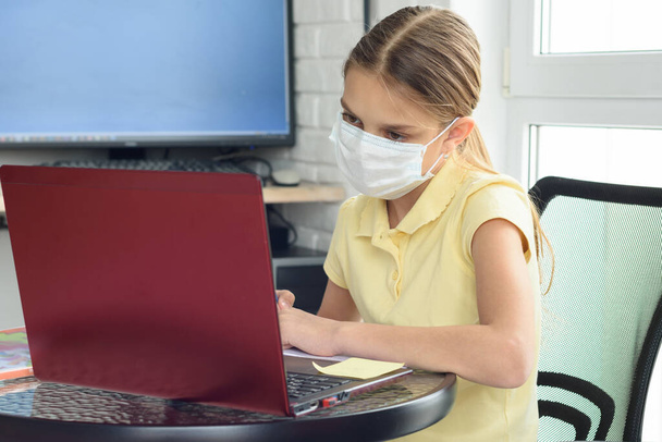 Ένα κορίτσι με ιατρική προστατευτική μάσκα υποβάλλεται σε εξ αποστάσεως εκπαίδευση - Φωτογραφία, εικόνα