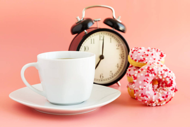 Weiße Tasse auf einer Untertasse mit schwarzem Kaffee, neben einem schwarzen Retro-Wecker und Donuts auf rosa Hintergrund. Frühstückszeit. Platz für Text. - Foto, Bild