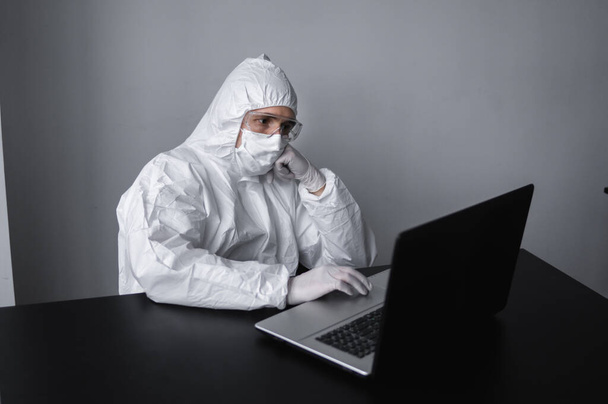 Man in medical mask and protective suit, rubber gloves sits at home and works at laptop at the table during quarantine. Designer, artist, architect, businessman at remote work in a pandemic covid. - Zdjęcie, obraz