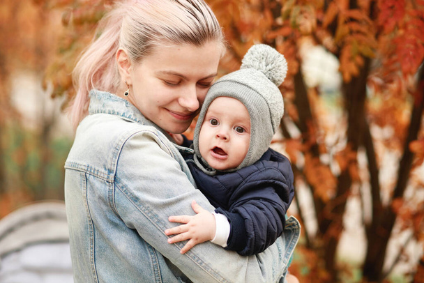 A stylish young mother with a small son in her arms walks in the autumn. Green grass and yellow leaves. - Фото, изображение