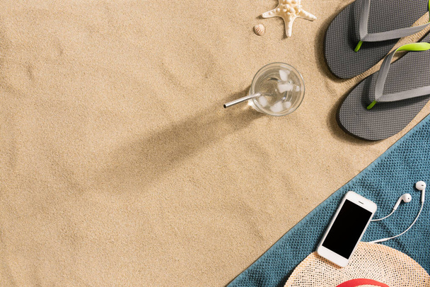 Летние каникулы композиция. Шлепанцы, шляпа, смартфон и стакан воды на песчаном фоне. Концепция туристического отдыха. Летний фон. Состав границы из полотенца
 - Фото, изображение