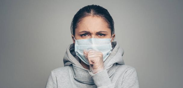Studio portrait of young woman wearing a face mask and gloves coughs, looking at camera, on gray background. Coronavirus pandemic, dust allergy, protection against virus concept panorama - Fotoğraf, Görsel