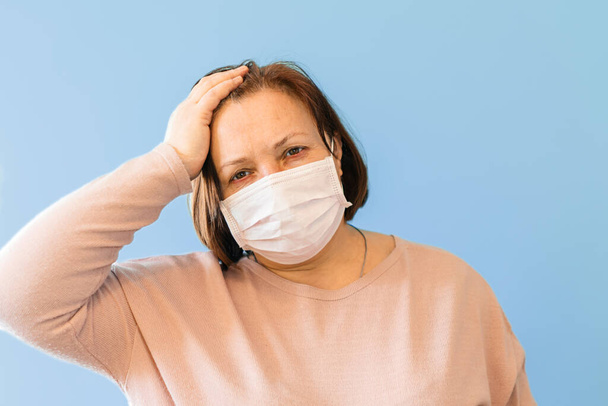 Unwell sick woman with surgical medical mask holding her head and looking sad, indoor, studio shot, isolated on blue background, covid-19 - Photo, Image