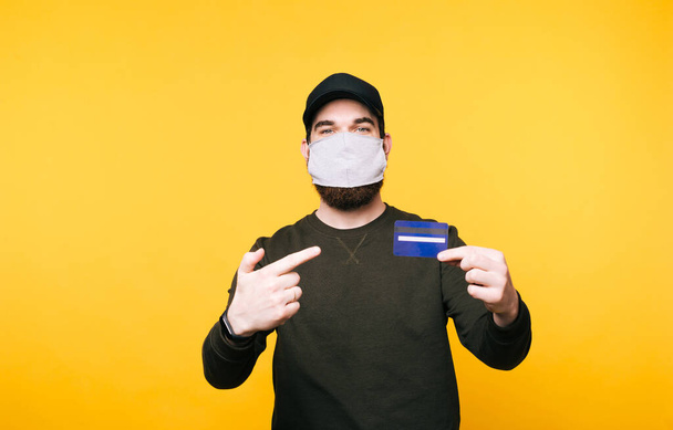 young man with facial mask pointing at credit card over yellow background - Photo, Image