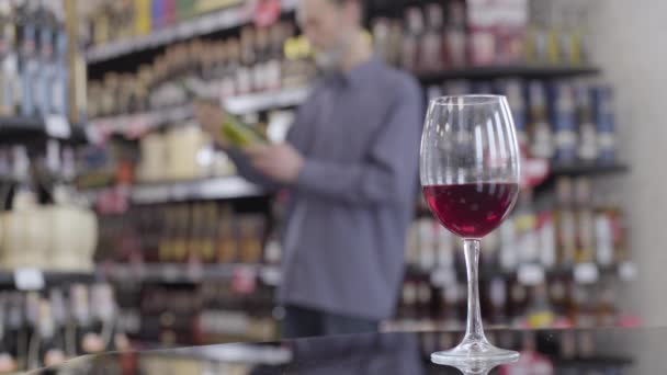 Close-up of wineglass standing on the table with blurred Caucasian man examining bottle of white wine at the background. Alcohol industry, liquor shop, lifestyle, degustation. - Filmagem, Vídeo