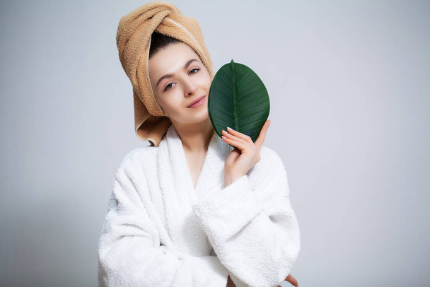 Beautiful woman in a white coat with a towel on her head and clear skin holds a green leaf - Fotó, kép