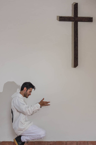 Hombre vestido de blanco rezando junto a una cruz de madera en fondo blanco
 - Foto, Imagen