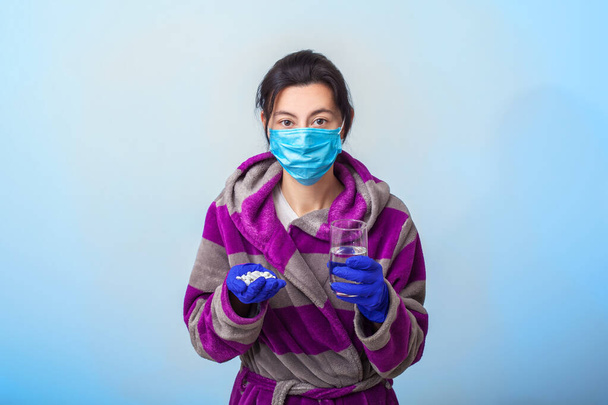 Koruyucu tıbbi maskeli ve eldivenli bir kız avuçlarında bir avuç hap ve bir bardak su tutuyor. Açık mavi arkaplan. Solunum maskesiyle. Coronavirus ve covid-19 konsepti. - Fotoğraf, Görsel