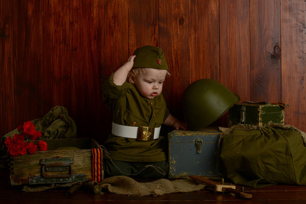 on the Victory Day on May 9, 1941-1945, a little boy soldier in uniform is sitting on old boxes, retro military atmosphere - Zdjęcie, obraz