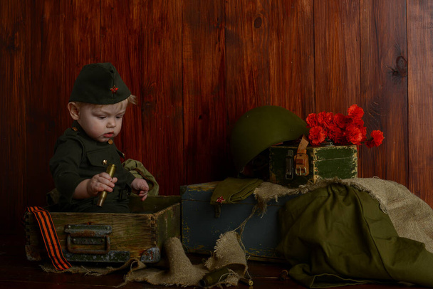 on the Victory Day on May 9, 1941-1945, a little boy soldier in uniform is sitting on old boxes, retro military atmosphere - Fotografie, Obrázek