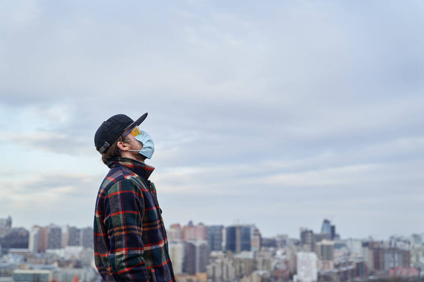 Jonge hipster man met masker om hem te beschermen tegen het corona virus, hopelijk kijkend naar een blauwe lucht op de achtergrond. Covid-19-viruspandemie of verontreinigingsconcept - Foto, afbeelding