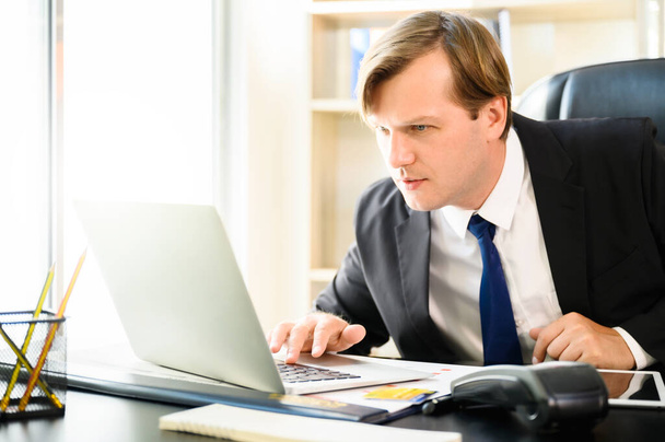 Молодой бизнесмен занят работой в офисе. Он использует ноутбук на столе в офисе - Фото, изображение