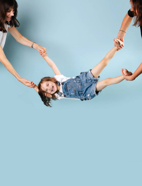 Two girls play with the child, hold the girl-toddler in a denim jumpsuit by the legs and hands and rock, the child rejoices and laughs. Isolated on a blue background - Photo, image