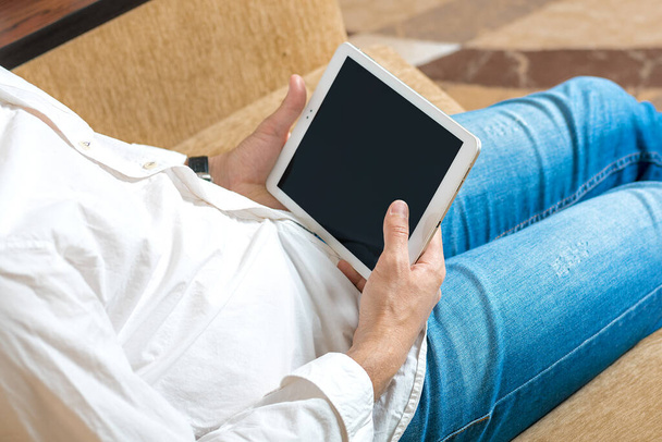 mladý muž v bílé košili a modré džíny pracovní a při pohledu na tablet s černou obrazovkou v domácí kanceláři na útulné béžové pohovce Zůstaňte doma, on-line práce, distanční učení koncept Top view Vysmát - Fotografie, Obrázek