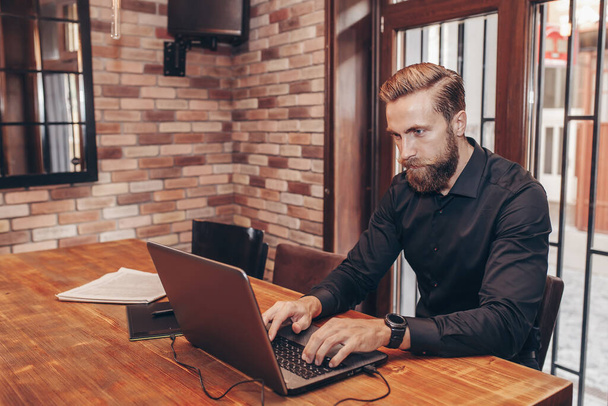 Νεαρός επιτυχημένος επιχειρηματίας που εργάζεται σε ένα φορητό υπολογιστή, ενώ κάθεται σε καφετέρια. - Φωτογραφία, εικόνα
