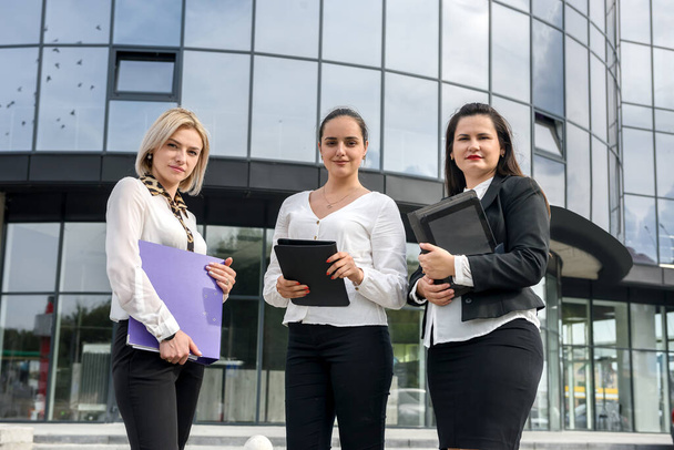 Équipe d'affaires. Trois femmes en costume avec des dossiers et une tablette posant à l'extérieur du bâtiment
 - Photo, image