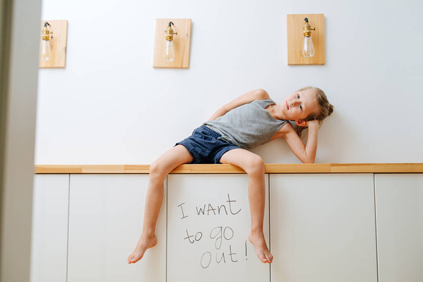 Little boy sitting on a corridor shelf over a board saying that he want's to go outside. Bored and tired of sitting at home during covid-19 self isolation. - Foto, imagen