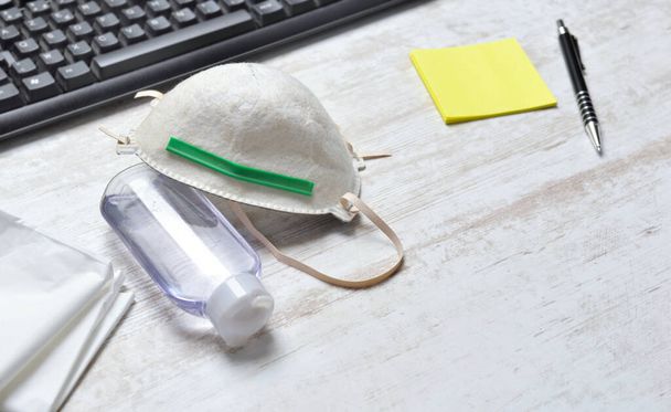 máscara de polvo para proteger la cara y la botella de desinfectante de manos en un escritorio junto al teclado y papel amarillo con un bolígrafo
  - Foto, Imagen