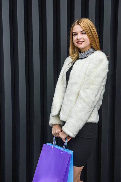 Stylish young woman in white fur coat and with colored bags on a background of black and white wall. Beauty and fashion. - Photo, Image