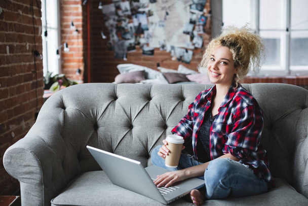 Young beautiful woman with curly hair with a laptop on the sofa at home searching the Internet. Freelancer. - Zdjęcie, obraz
