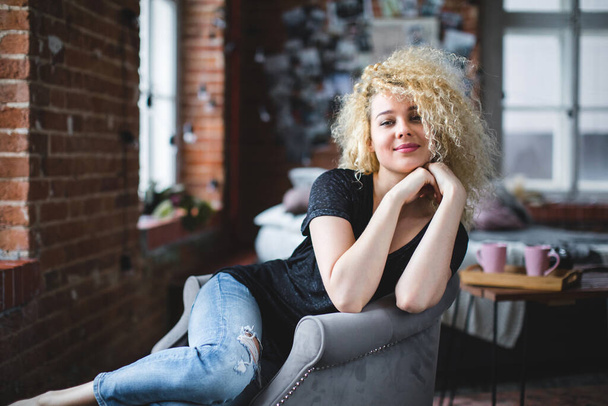 Woman with curly hair sitting in an armchair in a room with brick walls. - Fotoğraf, Görsel