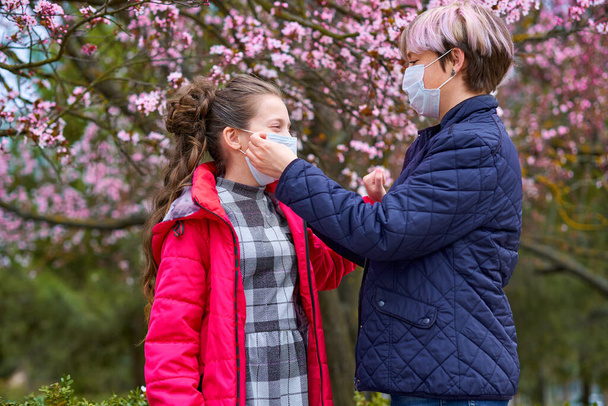 mother and daughter with a face mask are in the city outdoor, blooming trees, spring season, flowering time - concept of allergies and health protection from dusty air - Photo, Image