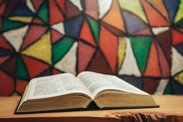 Zamknij otwartą Biblię na czerwonym drewnianym stole. Piękne witraże z tyłu. Pojęcie religii. - Zdjęcie, obraz