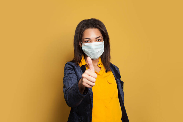 Mujer negra inteligente con máscara médica mostrando el pulgar hacia arriba sobre fondo amarillo
 - Foto, Imagen