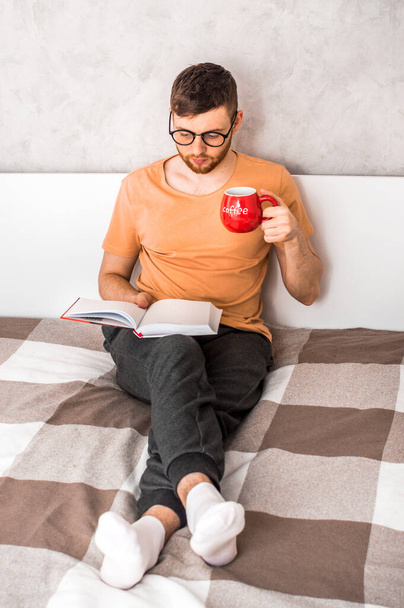 若い男がベッドで本を読み、コーヒーを飲む。垂直写真 - 写真・画像