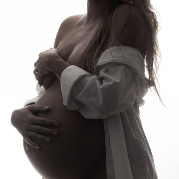Closeup pregnant woman in shirt holding her belly while standing over white background. Maternity concept - Fotografie, Obrázek