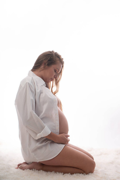 Mulher muito grávida em camisa branca e calcinha segurando sua barriga enquanto se ajoelha sobre fundo branco. Conceito de maternidade
 - Foto, Imagem