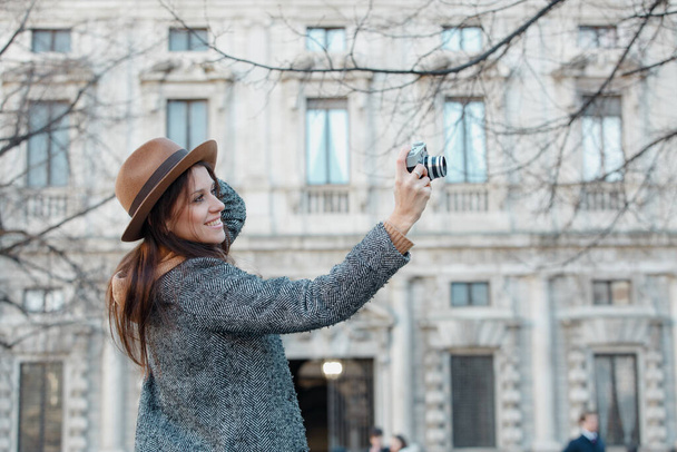 A young brunette girl takes pictures holding a hat with her hand. Against the background of a gray old building. - Foto, afbeelding