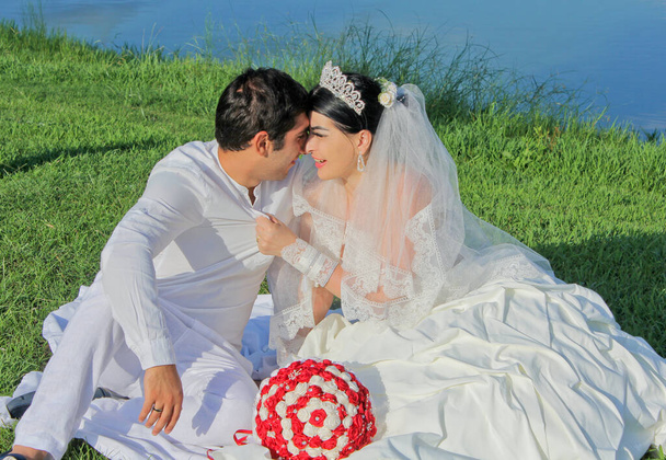 Funny couple of newlyweds on the green grass. Bride with a red bouquet in a wedding dress at the lake on background. Happy wedding day. Concept marriage, just married, family, honeymoon. - Photo, Image