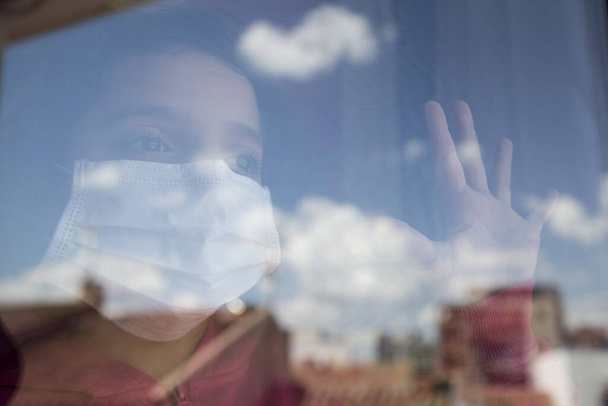 Hausgemachtes Mädchen mit Maske, das nach außen aus dem Fenster schaut. Mit der Stadt und den Wolken, die sich im Glas spiegeln. Mit der Hand am Fenster sehnt sie sich zurück auf die Straße. Konzept covid-19. - Foto, Bild
