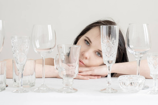 Young woman with a different wine glasses on the table on the white background. A lot of glasses for drinks with sunlight and shadow. - Foto, Imagen