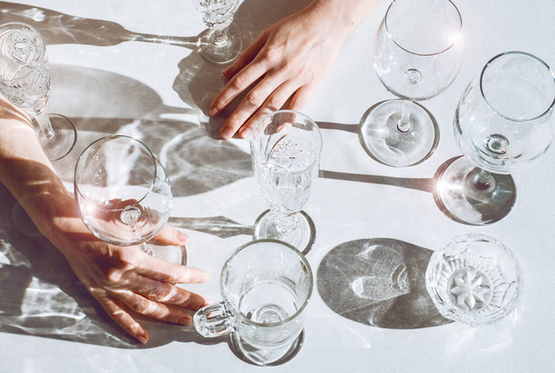 Manos de mujer con diferentes copas de vino sobre la mesa sobre fondo blanco. Una gran cantidad de vasos para bebidas con luz solar y sombra
. - Foto, imagen