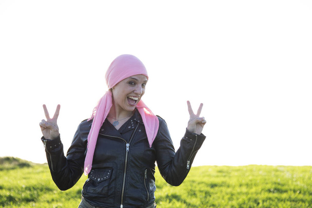 Γυναίκα με μαύρο μπουφάν και ροζ μαντίλα σε χωράφι. Με δάχτυλα σε σχήμα νίκης. Έννοια του καρκίνου του μαστού. - Φωτογραφία, εικόνα