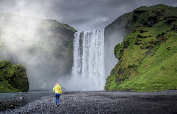 Молодая девушка, идущая рядом с водопадом Скогафосс летом, Исландия
 - Фото, изображение
