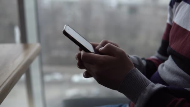 Een jongeman met een telefoon close-up. Een man zit in een café bij het panoramische raam met een smartphone in zijn handen. Zicht vanuit het raam. - Video