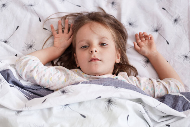 Image of sleepy sweet beautiful little girl lying in her bed, raising her arms, looking directly at camera, having serious facial expression, wearing sleepwear, waking up in morning. Sleep concept. - Фото, зображення