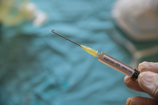 Inyección de cerca de una jeringa con la palabra vacuna contra el coronavirus.La mano que la sostiene presiona el émbolo y sale una gota de la aguja. En el fondo borroso una máscara y el gel desinfectante
 - Foto, Imagen
