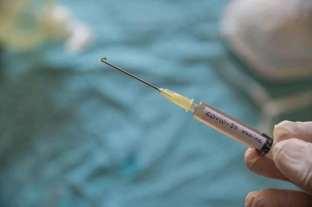 Крупный план шприца со словом коронавирусная вакцина. Рука, держащая его, нажимает вантуз и капля выходит из иглы. На размытом фоне маска и гель для дезинфекции
 - Фото, изображение
